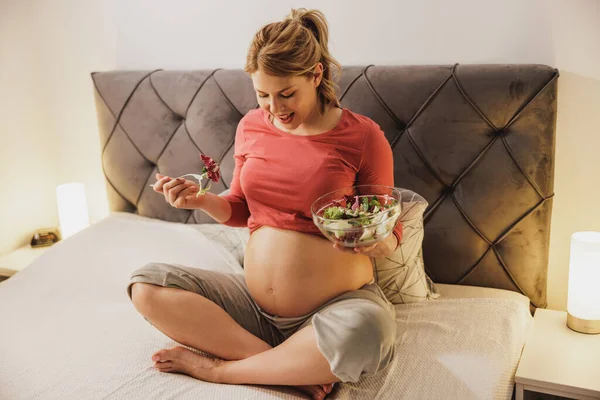 幸せな妊娠中の女性は ベッドルームのベッドに座って新鮮な野菜サラダを食べています — ストック写真