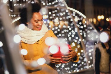 Orta yaşlı siyahi bir kadın, şehirde bir şenlik gecesinde açık hava Noel pazarında hediyesini tutuyor..