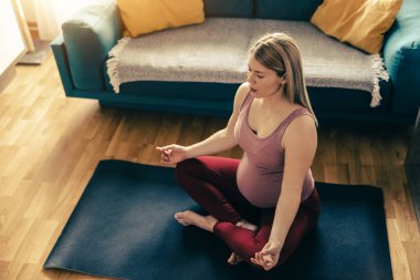 Genç hamile kadın sabahları evde yoga yapıyor. Oturma odasındaki paspasın üstünde egzersiz yapıyor..