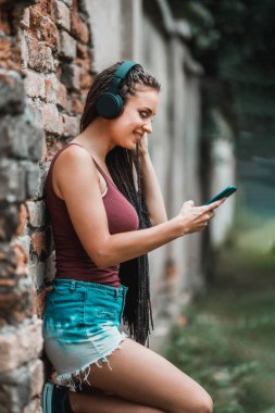 Tuğla duvara karşı müzik dinlerken akıllı telefon kullanan uzun saçlı hoş bir genç kadın..