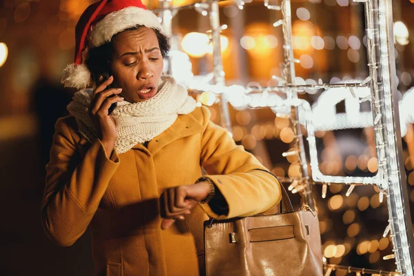 Middle age worried black woman looking at her watch while talking phone on the festive Christmas market in the city.