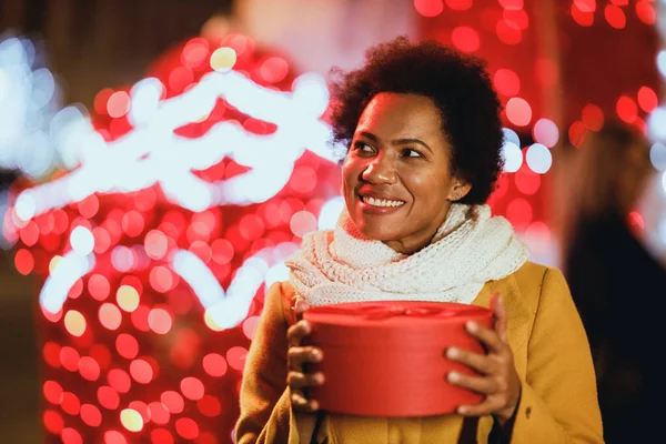 Smiling black woman holding gift on festive Christmas market outdoors in winter night at the city.
