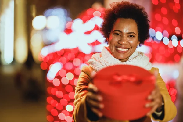 Smiling black woman holding gift on festive Christmas market outdoors in winter night at the city.