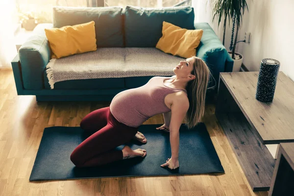 A pregnant woman in sports clothing is stretching and exercising mobility to promote wellbeing in her living room.
