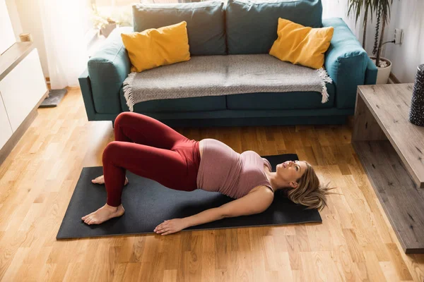 Young pregnant woman exercising at home in the morning. She is doing mobility exercise in her living room.
