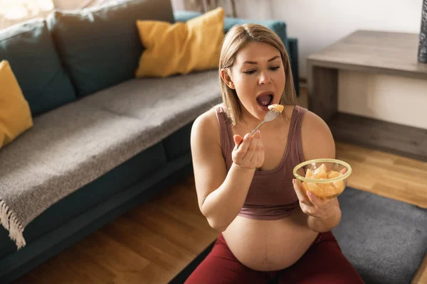 自宅で運動しながら新鮮なフルーツサラダを食べている幸せな若い妊婦 彼女は朝のリビングルームで運動マットでリラックス — ストック写真