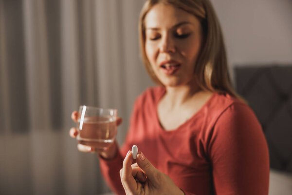 Pregnant woman holding glass with water and drinking vitamin pill in the morning.