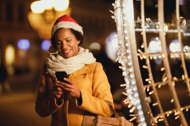 Orta yaşlı Afrikalı bir kadın şehirdeki şenlikli bir gecede Noel pazarında akıllı telefonuyla yazı yazıyor..