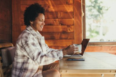 Siyah bir kadın sabah çayı içip dizüstü bilgisayar kullanırken, tatilini rahat, ahşap bir evde geçiriyor..
