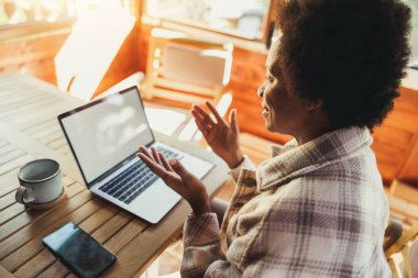 Siyah bir kadın sabahları bilgisayarında video görüşmesi yapıyor. Tatil zamanını ahşap bir evde geçiriyor..