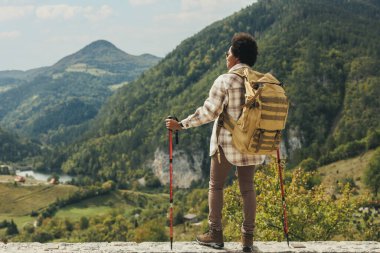 Sırt çantası ve yürüyüş direkleri olan siyah bir kadının arka görüntüsü doğada duruyor ve dağda yürüyüş yaparken temiz havanın tadını çıkarıyor..