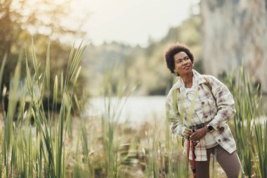 Sırt çantalı olgun siyahi kadın nehir kenarında duruyor doğa yürüyüşünden dolayı dinleniyor..