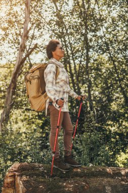 Sırt çantası ve yürüyüş direkleri olan olgun siyah bir kadın güzel bir sonbahar gününde dağlarda yürüyüş yapıyor..