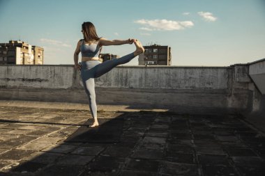 Young woman practicing yoga while doing workout outdoors on a rooftop terrace.