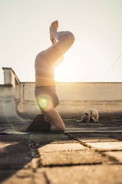 Relaxed woman practicing yoga headstand on a rooftop terrace at sunset.