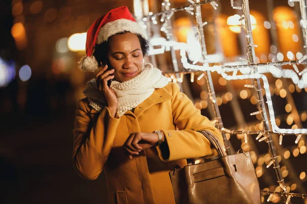 Middle age black woman looking at her watch and talking phone on the festive Christmas market in the city.