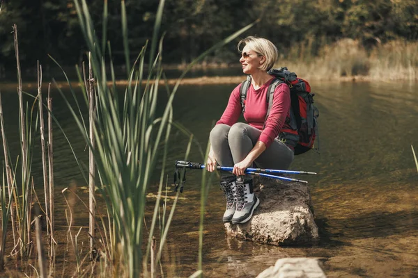 stock image Mature woman with backpack sits alone on a rock and enjoying the view of lake in mountains.