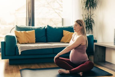 Evde yoga yapan hamile bir kadın. Egzersiz paspası üzerinde çalışıyor ve oturma odasında esniyor..