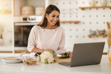 Evde sağlıklı bir yemek hazırlarken bilgisayarı kullanarak biriyle görüntülü konuşma yapan genç bir kadının fotoğrafı..