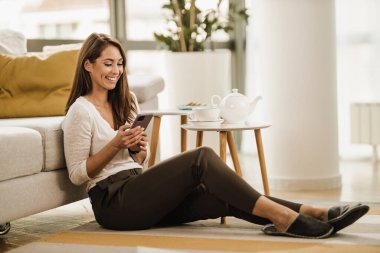 Oturma odasında yerde oturan ve akıllı telefon kullanan genç bir kadını rahatlatıyor. Sosyal medyada boş vakitleri var..