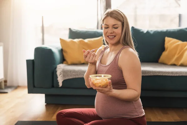 满意地期待着女人在家里锻炼的时候吃新鲜的水果沙拉 早上她在客厅的练习垫上放松 — 图库照片