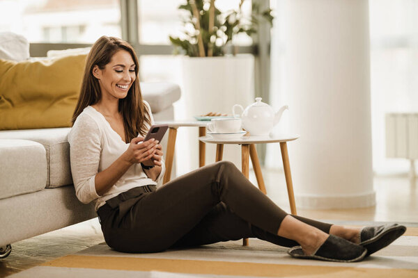 Relaxing young woman sitting on the floor in her living room and using a smartphone. She having leisure time surfing social media.