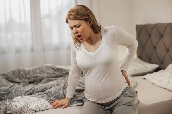 stock image Worried pregnant woman holding her back in pain while sitting on a bed in bedroom.