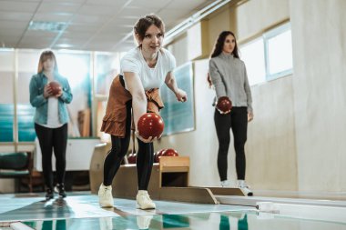 A cute girl throwing the bowling ball while her friends are standing in the back.