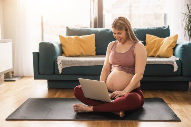 Genç hamile kadın sabah oturma odasında dinlenirken dizüstü bilgisayar kullanıyor..