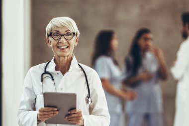 A successful senior female doctor looking at camera and holding digital tablet in a hospital hallway.