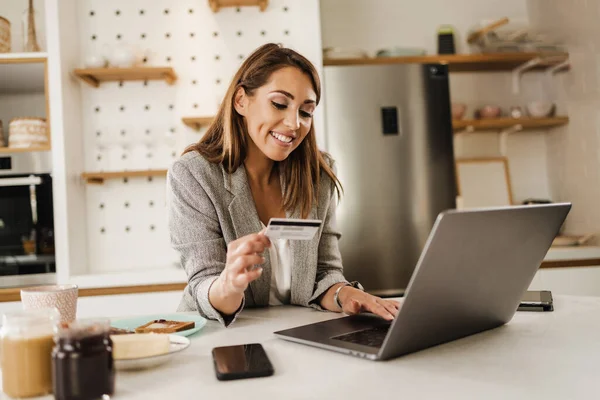 Multi-tasking young businesswoman using laptop and paying bills with credit card in her kitchen at the morning while getting ready to go to work.