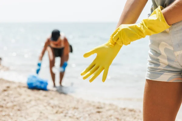 stock image Close-up of a unrecognizable female volunteer putting protective rubber yellow gloves while cleaning sea beach area from the trash.