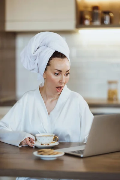 穿着浴衣的年轻女人在家里喝咖啡的时候在笔记本电脑上冲浪 — 图库照片