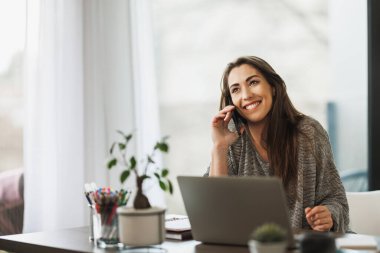 Evde dizüstü bilgisayarıyla çalışan ve akıllı telefonuyla konuşan mutlu genç bir kadın..