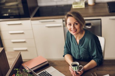 Olgun bir bayan serbest yazar ev ofisinde çalışırken bir fincan kahvenin tadını çıkarıyor. Kameraya bakıyorum..