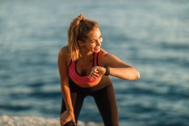 Mutlu genç bayan koşucu deniz kıyısındaki eğitimden sonra dinleniyor, kalp atışlarına bakıyor ve akıllı yamağındaki gelişmeleri kontrol ediyor..