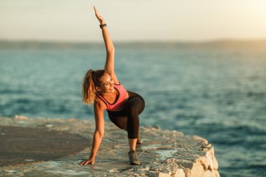 Mutlu genç bayan koşucu deniz kıyısındaki eğitim sırasında ısınma egzersizleri yapıyor..