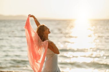Çekici genç bir kadın yaz tatilinde eğleniyor ve eğleniyor. Plajda elinde şeffaf bir eşarpla poz veriyor..