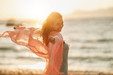 Çekici genç bir kadın yaz tatilinde eğleniyor ve eğleniyor. Plajda elinde şeffaf bir eşarpla poz veriyor..