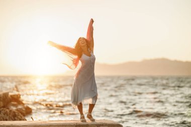 Çekici genç bir kadın yaz tatilinde eğleniyor ve eğleniyor. Plajda elinde şeffaf bir eşarpla poz veriyor..