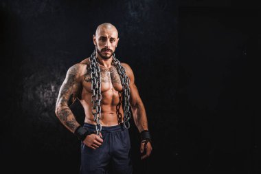 Genç bir fitness adamı salonda duruyor ve ağır bir zincirle antrenman yapmaya hazırlanıyor. Kameraya bakıyorum..