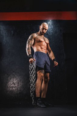 Genç bir fitness adamı salonda duruyor ve ağır bir zincirle antrenman yapmaya hazırlanıyor..
