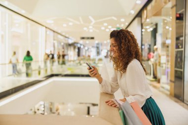 Alışveriş sonrası çantalarını taşıyan akıllı telefonunu kullanan genç bir kadının fotoğrafı..