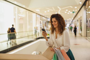 Cep telefonuyla mesaj atan ve alışveriş merkezinde poşet tutarken gülümseyen çok mutlu bir kadının portresi..