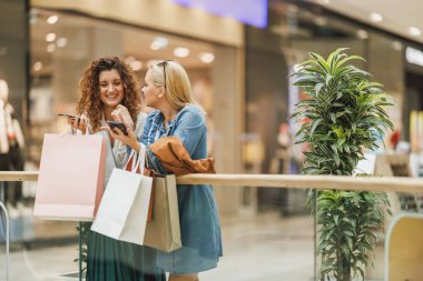 Akıllı telefon kullanan ve alışveriş merkezinde alışveriş yaparken ayakta duran iki güzel ve gülümseyen kadının fotoğrafı..
