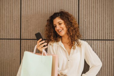 Alışveriş çantalarını tutarken akıllı telefonuyla mesaj atan çok mutlu bir kadının bir alışveriş merkezinin arka planında duvar resmi..