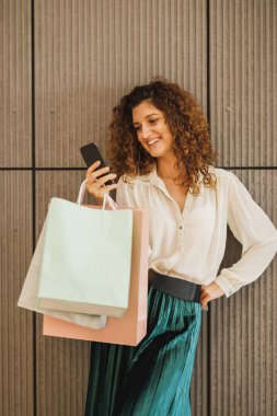 Alışveriş merkezinin arka planında akıllı telefonunu duvara dayayıp alışveriş torbaları tutan bir kadının fotoğrafı..