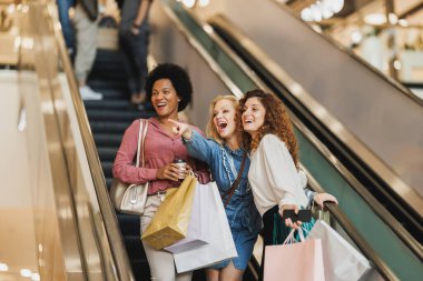 Çekici bir kadın alışveriş merkezinde en iyi arkadaşlarıyla yürüyen merdivende dikilirken bir şeyi işaret ediyor..