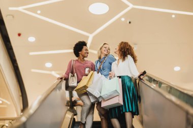 Alışveriş merkezinde alışveriş yaparken elinde poşetlerle yürüyen merdivende gülümseyen üç çekici kadının fotoğrafı..