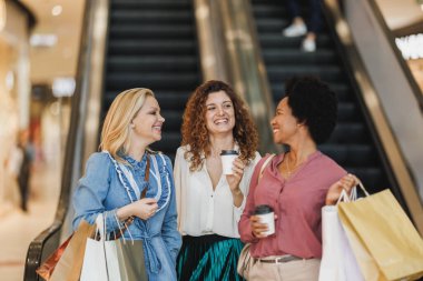 Kara Cuma ya da Siber Pazartesi satışları sırasında alışveriş merkezinde alışveriş torbalarıyla yürürken gülümseyen üç kız arkadaş konuşuyor..